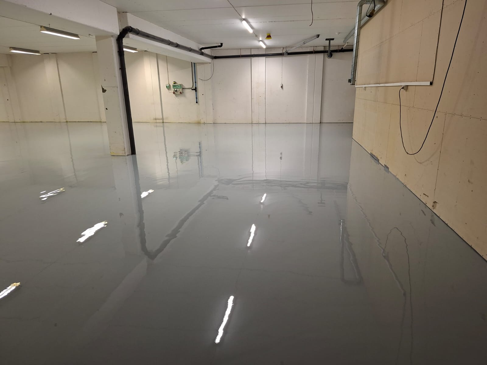 Błyszcząca, szara posadzka w hali magazynowej w Pile. Widoczne odbicia świetlówek w gładkiej powierzchni. Ściany w jasnych kolorach, instalacje pod sufitem.