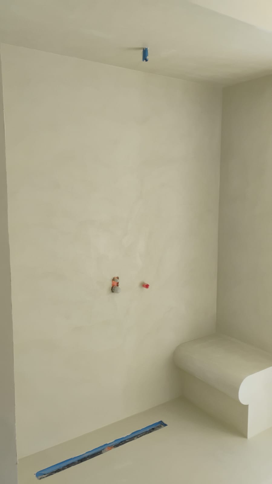 Jasne wnętrze łazienki po nałożeniu beżowej powłoki na ściany i podłogę, widoczne otwory na instalacje i zabezpieczony odpływ liniowy.