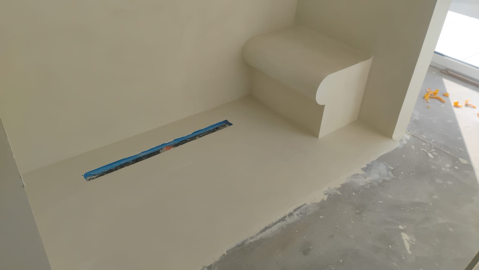 Beżowa, gładka powierzchnia w łazience z odpływem liniowym i półką. Widoczne niedoskonałości wykończenia przy podłodze. Minimalistyczny design.