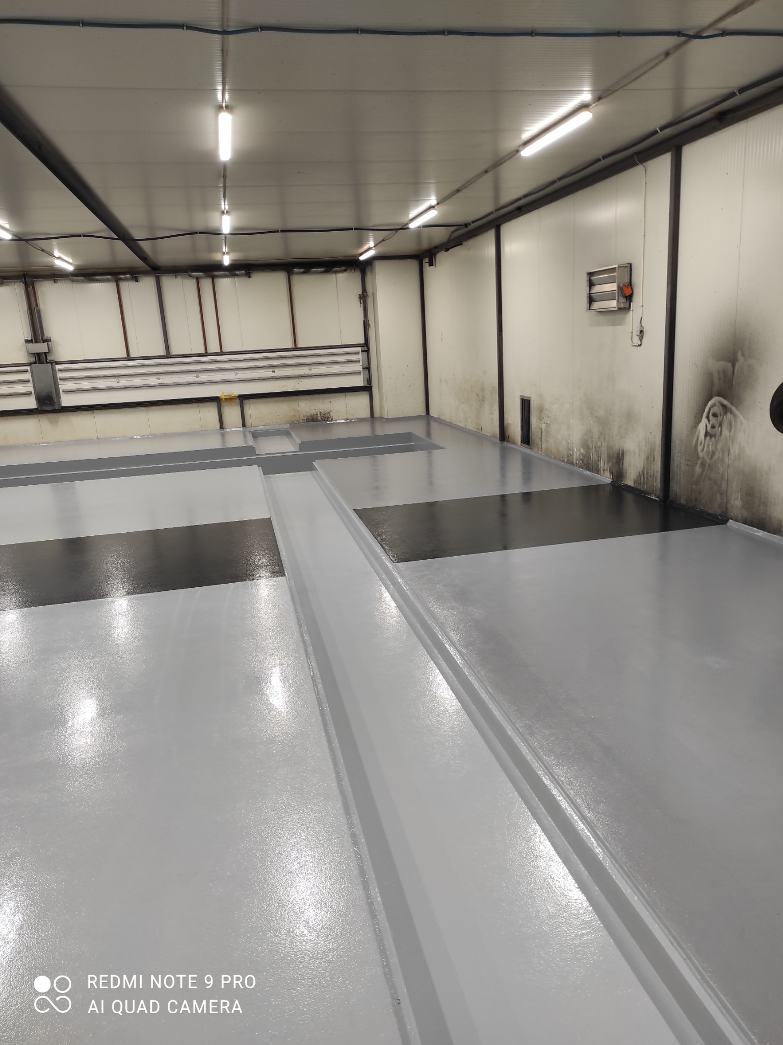 Jasnoszara, błyszcząca podłoga w hali przemysłowej z ciemniejszymi, prostokątnymi wstawkami. Widoczne elementy instalacji pod sufitem. Ściany białe, miejscami zabrudzone.