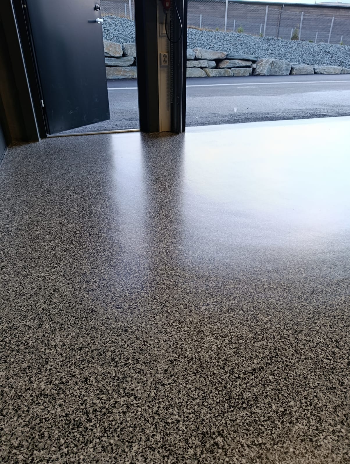 Błyszcząca, szara posadzka w garażu z otwartymi drzwiami. Widoczne drobne, ciemne i jasne plamki tworzące wzór. Na zewnątrz widoczny fragment drogi i kamienny murek.