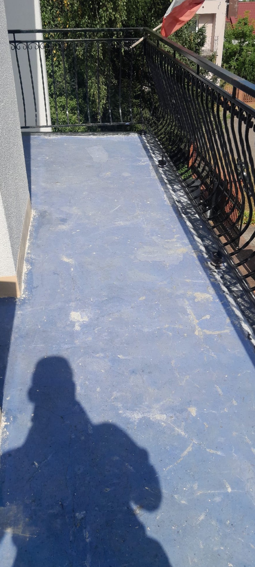 Niebieska posadzka żywiczna na balkonie z czarną, ozdobną balustradą. Widoczny cień osoby fotografującej. W tle zieleń drzew i fragment budynku.