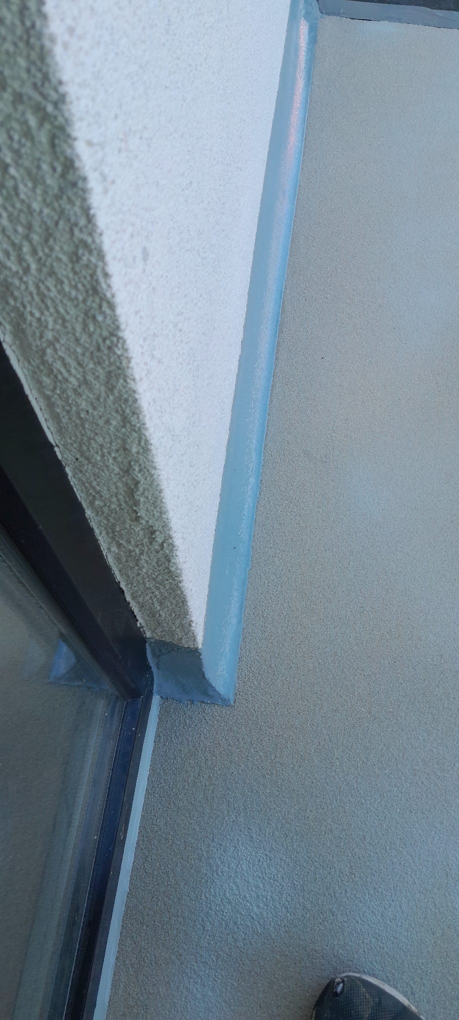 Szara, gładka powierzchnia podłogi przy ścianie z niebieskim wykończeniem, widok z góry. Fragment buta w rogu kadru.