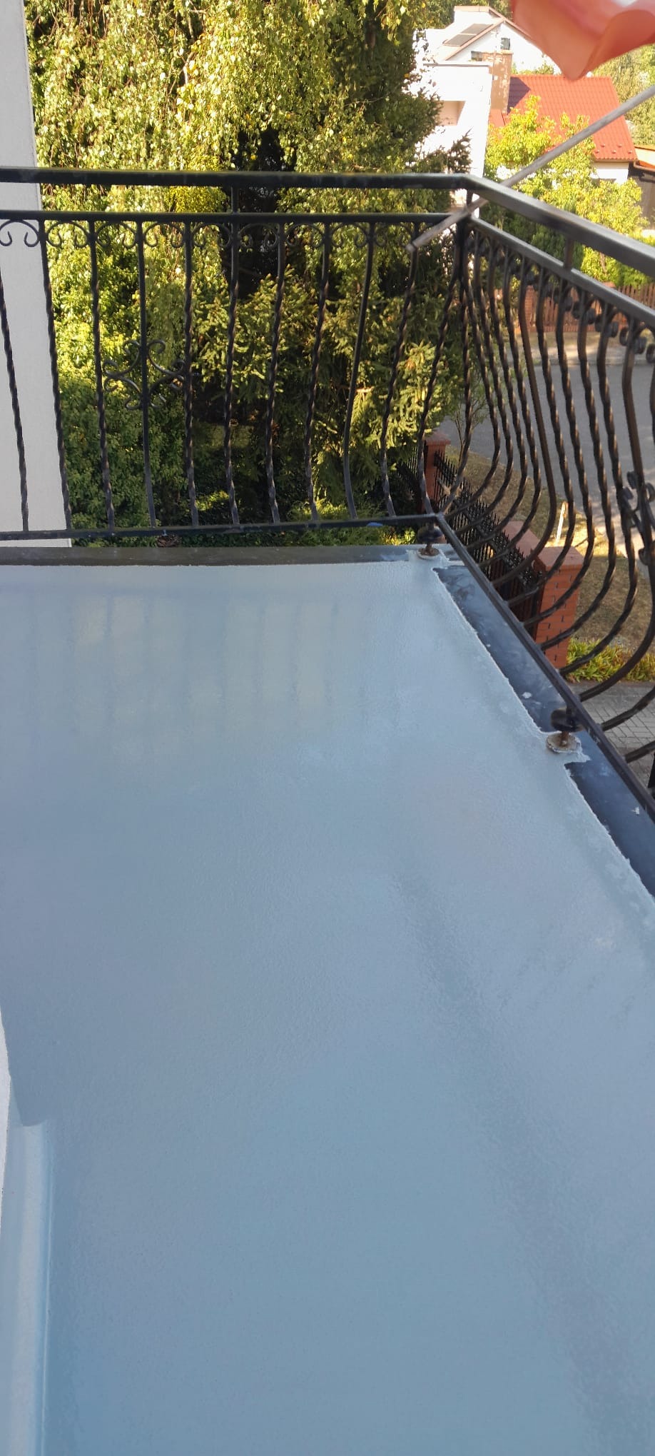 Błękitna, gładka powierzchnia balkonu po renowacji, widok z góry. Metalowa balustrada z ozdobnymi elementami. W tle zieleń drzew i zabudowania.