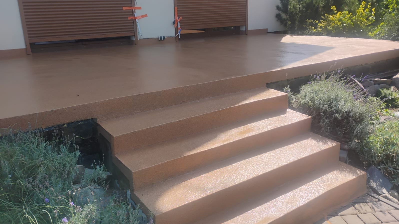 Beżowa, gładka posadzka na schodach zewnętrznych i tarasie, z widocznymi detalami wykończenia i roślinnością w tle. Całość w naturalnym świetle.