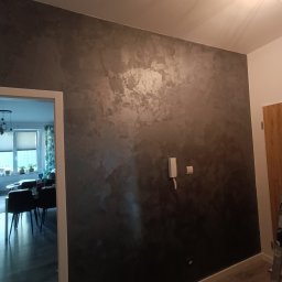 Laifdyb - Ściana w pokoju po dekoracyjnym wykończeniu, z efektem cieniowania w odcieniach szarości, widoczny fragment drabiny malarskiej i wiaderko po farbie w rogu pokoju.