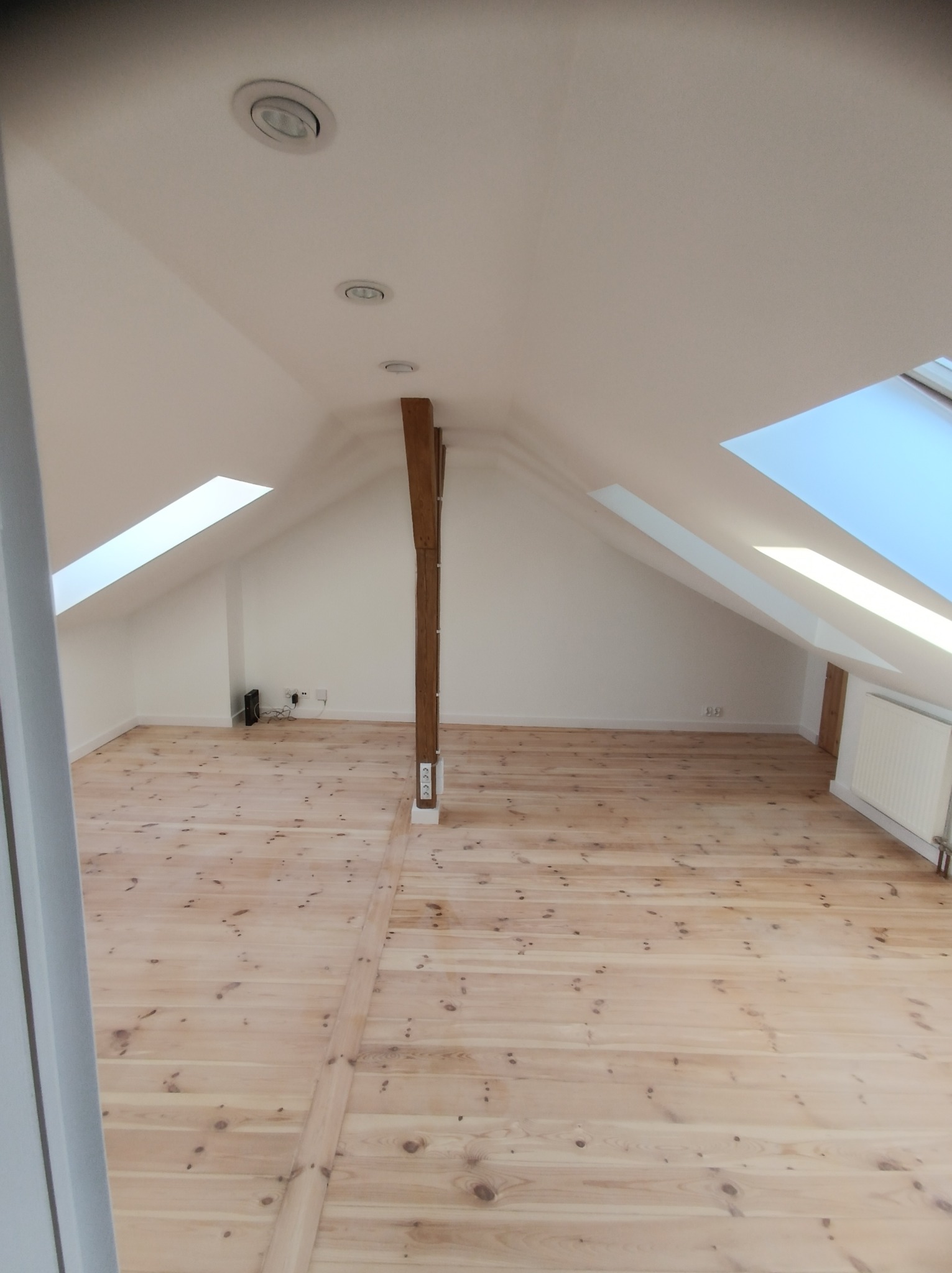 Poddasze po remoncie z drewnianą podłogą i białymi ścianami, widoczne okna dachowe i drewniany słup konstrukcyjny, oświetlenie punktowe w suficie.