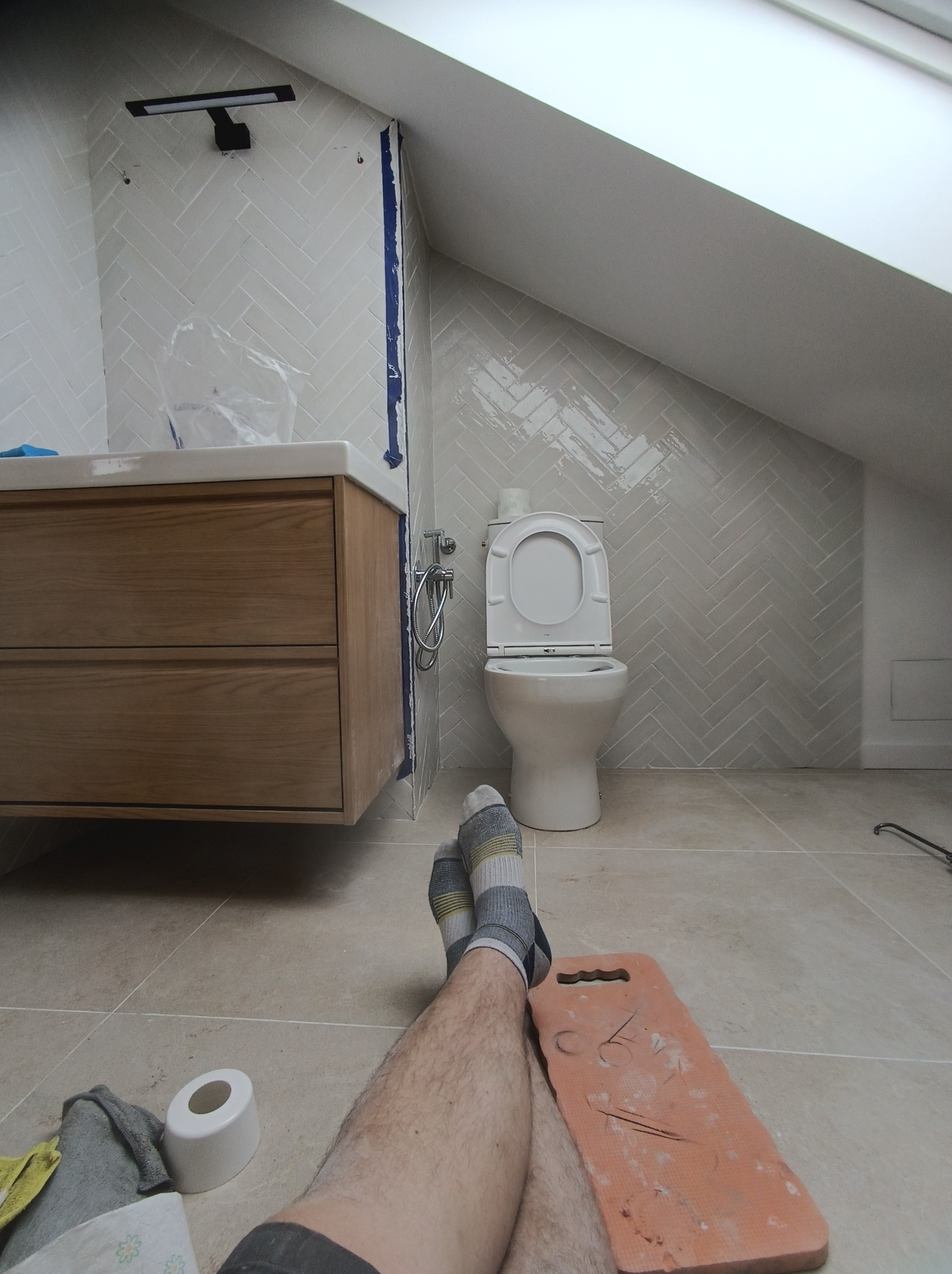 Ukończona łazienka na poddaszu z toaletą, umywalką z szafką z drewna i wykończonymi ścianami w jasnych płytkach ułożonych w jodełkę, widok z perspektywy osoby leżącej na podłodze w skarpetkach.