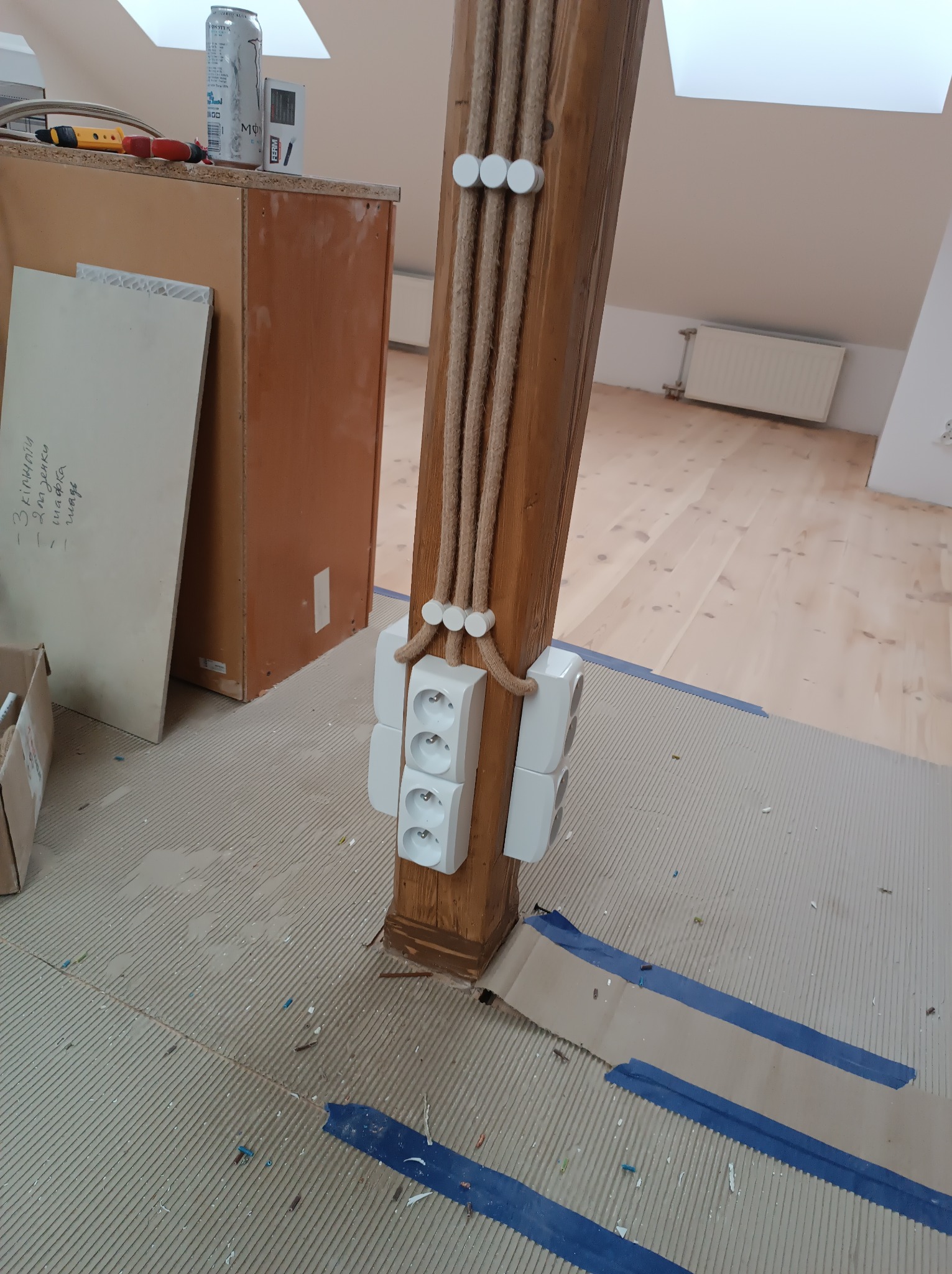 Drewniany słup z zamontowanymi gniazdkami elektrycznymi i poprowadzonymi kablami w ozdobnych oplotach, widoczne zabezpieczenia podłogi taśmą malarską, w tle poddasze z oknem dachowym.
