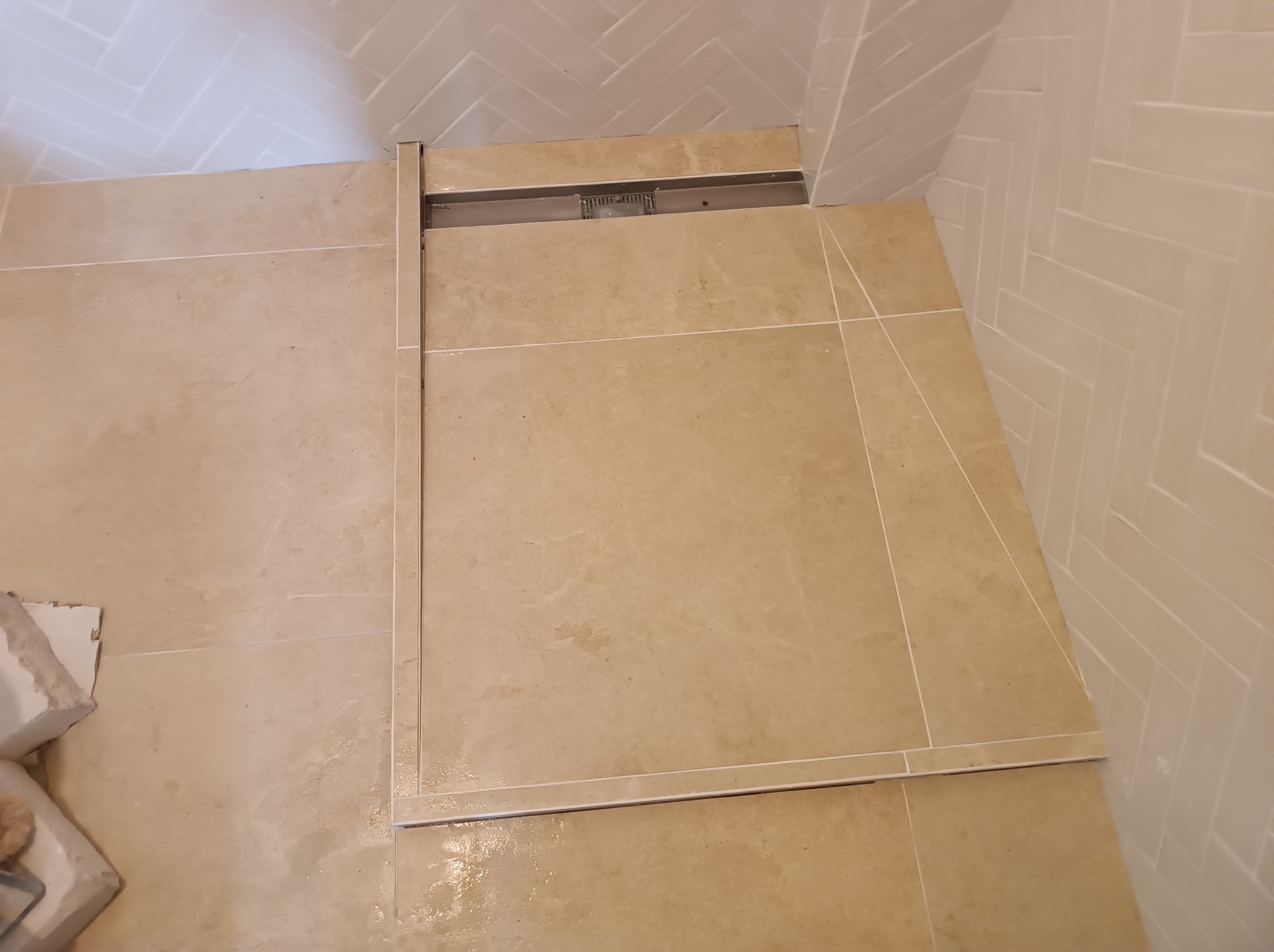 Beżowe płytki podłogowe z odpływem liniowym w kabinie prysznicowej, białe płytki ścienne ułożone w jodełkę, widoczne fragmenty zaprawy i płytek w rogu.
