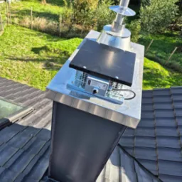 Widok z góry na komin z zamontowanym wentylatorem dachowym z czarną pokrywą, srebrną podstawą i metalowym wylotem, na tle zielonego trawnika i drzew.