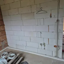 Wnętrze w trakcie prac elektrycznych: ściana z betonu komórkowego z wystającymi przewodami, puszki elektryczne, w tle widoczny fragment konstrukcji i drabina.