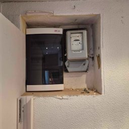 Highlander Energy Mateusz Góralczyk - Wnęka w ścianie z zamontowaną skrzynką bezpiecznikową ETI i licznikiem energii elektrycznej, widoczne ślady po wierceniu, surowa ściana z fakturą baranka.
