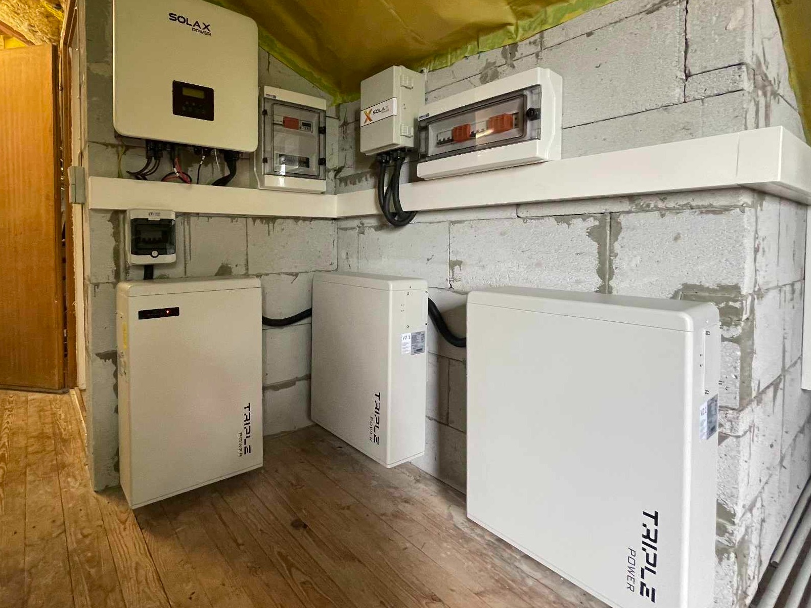 Białe akumulatory energii z logo Triple Power, falownik Solax i skrzynki elektryczne zamontowane na ścianie z bloczków betonowych w pomieszczeniu z drewnianą podłogą i folią na suficie.