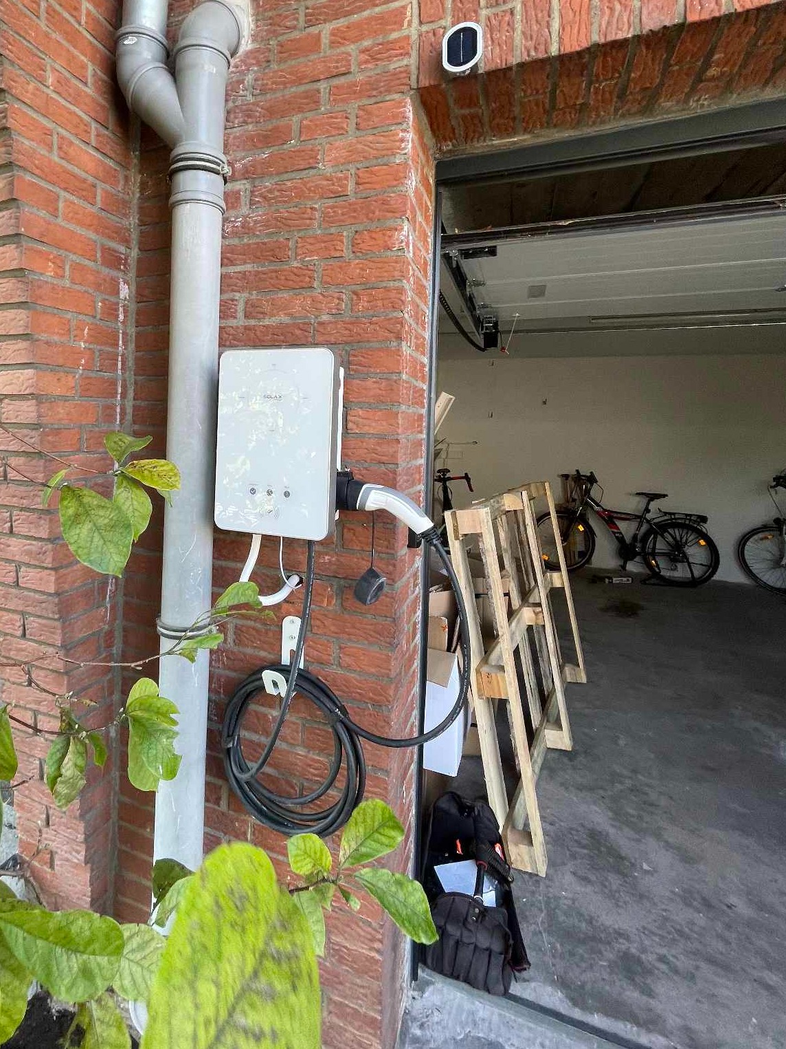 Biała stacja ładowania samochodu elektrycznego Solax zamontowana na ceglanej ścianie garażu, z widocznym kablem i wtyczką, oraz fragmentem roślinności w lewym dolnym rogu.