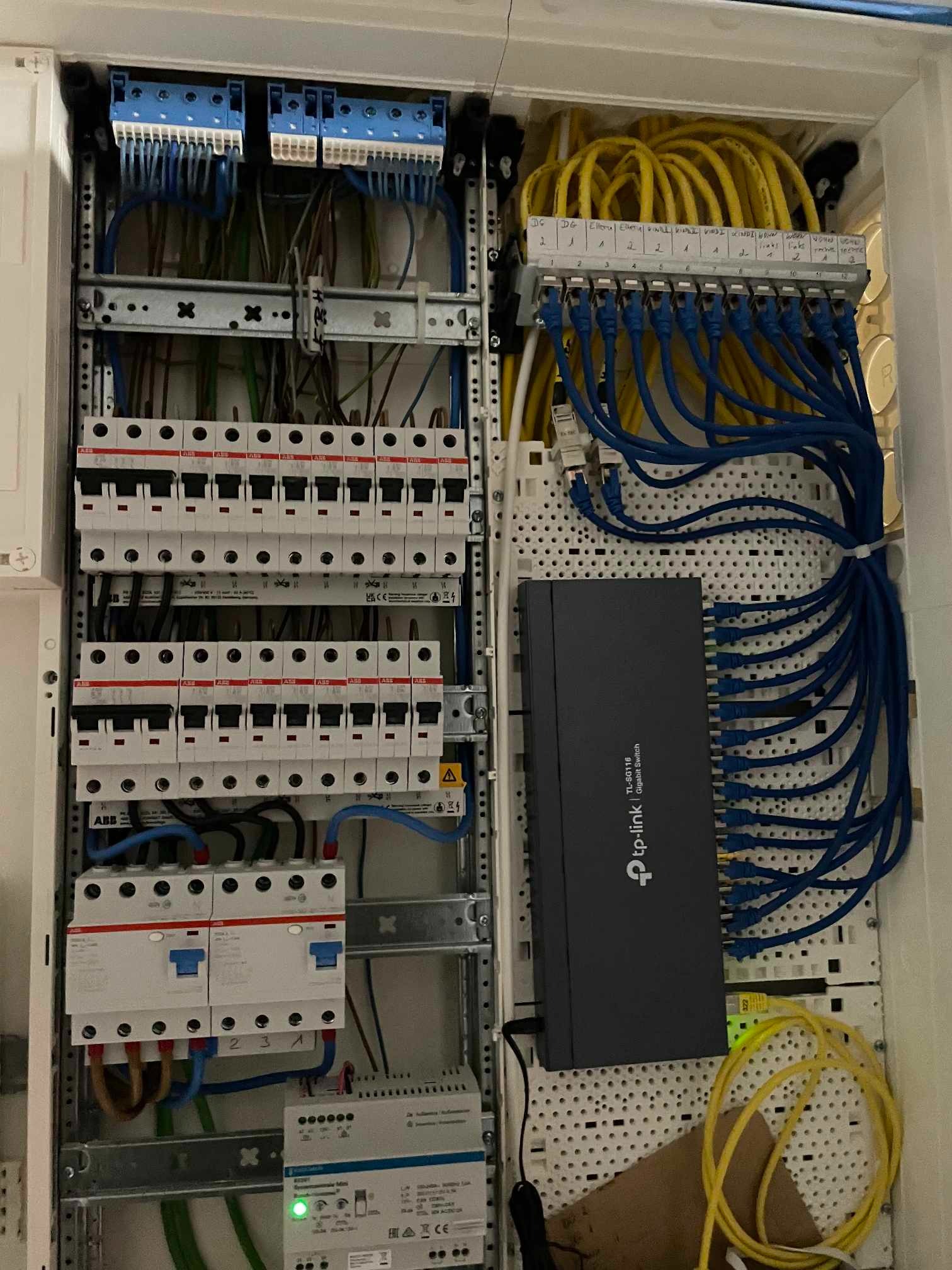 Rozdzielnica elektryczna z zabezpieczeniami, bezpiecznikami, przewodami i przełącznikiem sieciowym TP-Link, kable sieciowe w kolorach żółtym i niebieskim, widoczne oznaczenia na listwach zaciskowych.