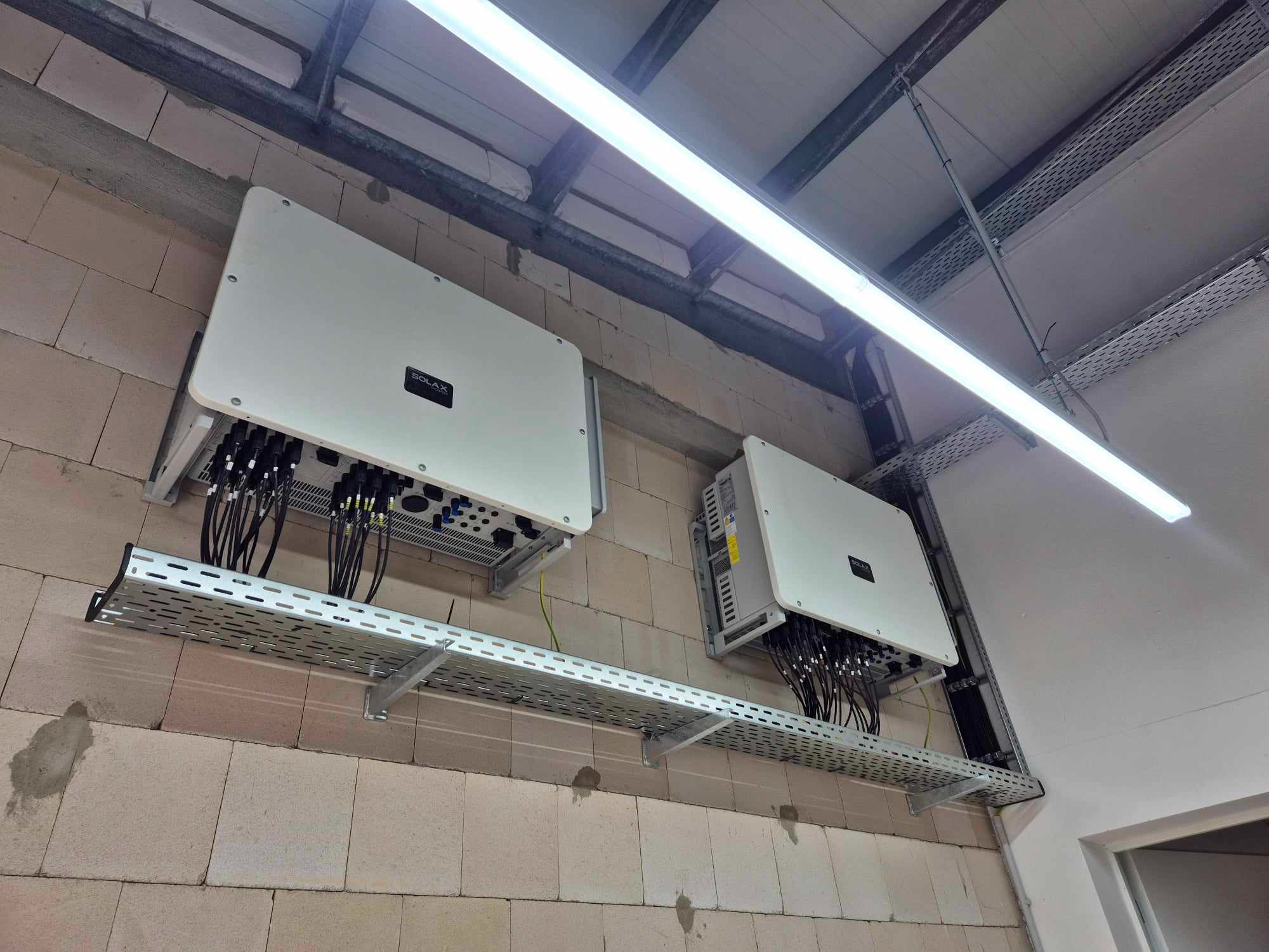 Dwa białe inwertery Solax zamontowane na perforowanych metalowych półkach na ścianie z bloczków, z widocznymi kablami i oświetleniem LED.