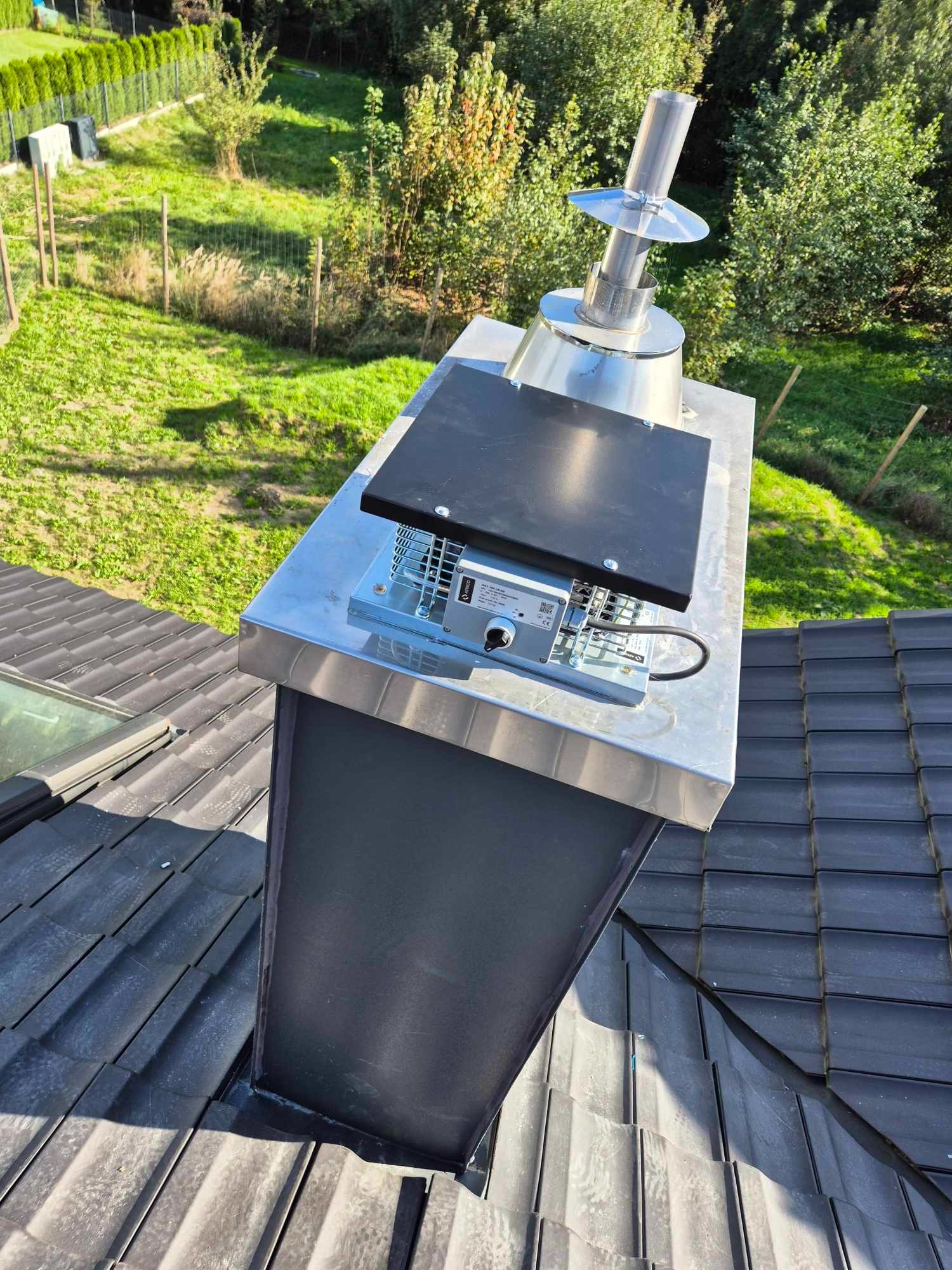 Widok z góry na komin z zamontowanym wentylatorem dachowym z czarną pokrywą, srebrną podstawą i metalowym wylotem, na tle zielonego trawnika i drzew.