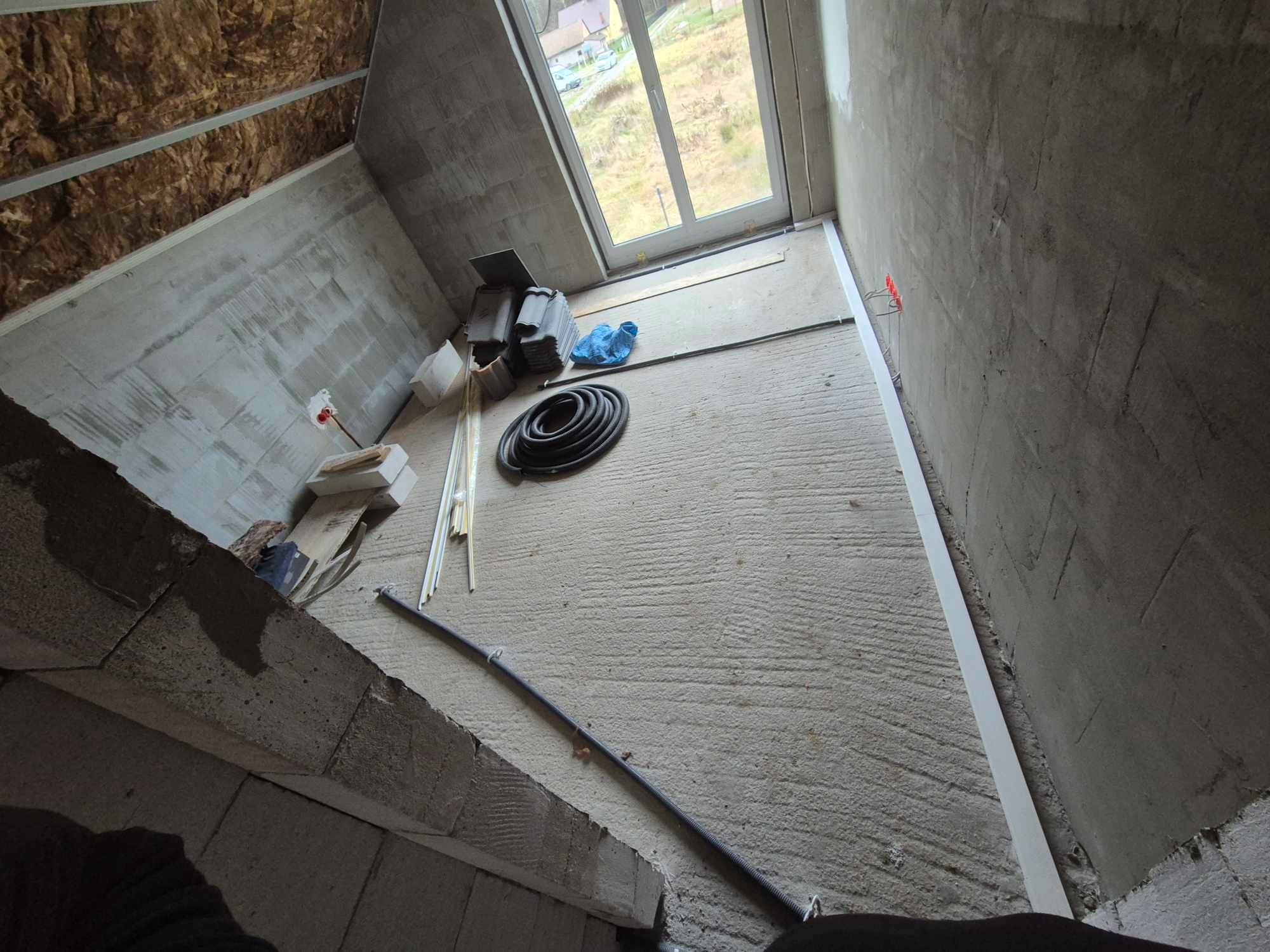 Surowy stan poddasza z oknem dachowym i przygotowanymi przewodami elektrycznymi, widoczne puszki instalacyjne w ścianie, wełna mineralna w skosach dachu oraz zwój kabla na podłodze.