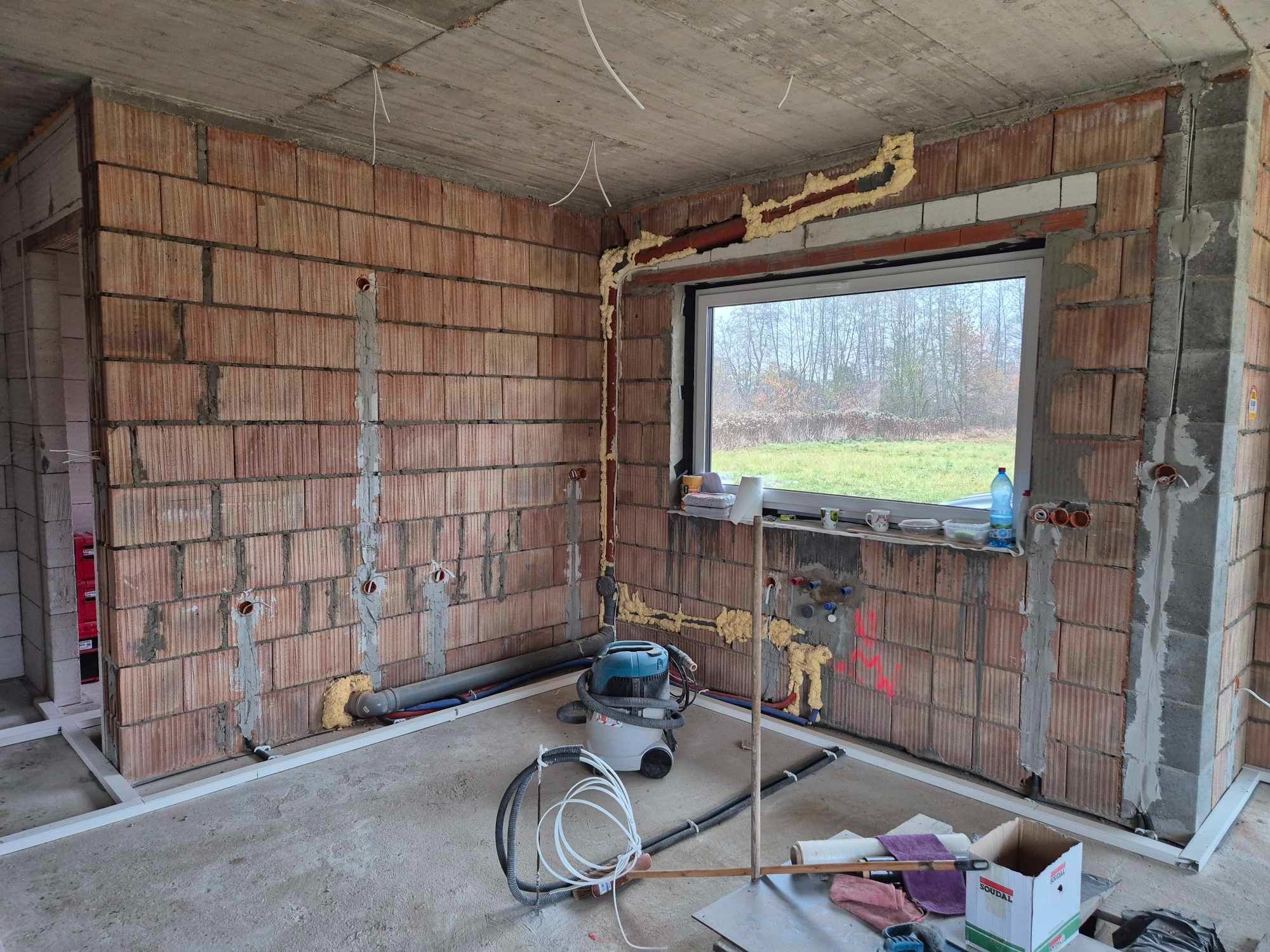 Wnętrze w stanie surowym z widocznymi przewodami elektrycznymi w ścianach, otwory pod gniazdka i puszki, pianka montażowa wokół okna, rury instalacyjne oraz odkurzacz przemysłowy na podłodze.
