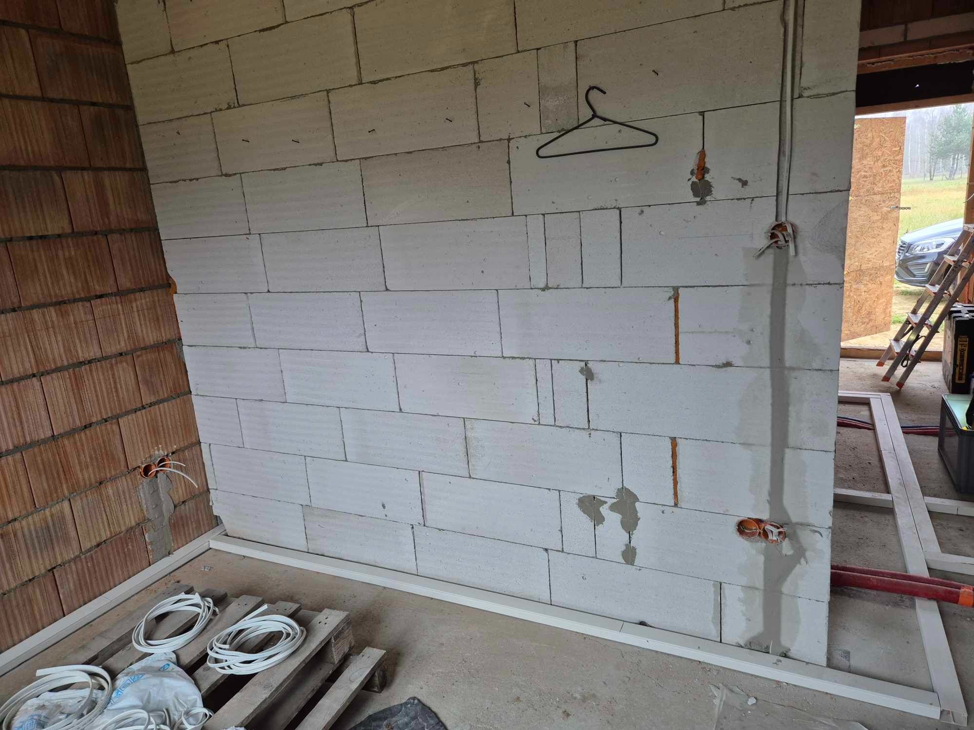 Wnętrze w trakcie prac elektrycznych: ściana z betonu komórkowego z wystającymi przewodami, puszki elektryczne, w tle widoczny fragment konstrukcji i drabina.