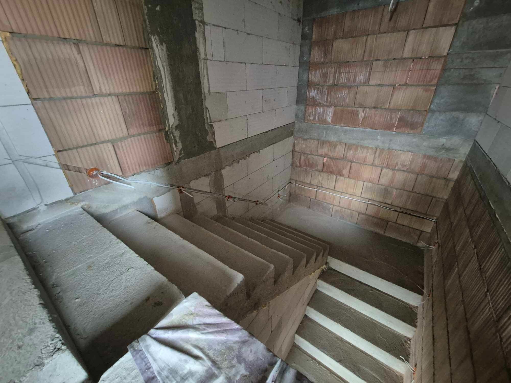 Surowe schody betonowe w trakcie budowy, widoczne przewody elektryczne wystające ze ściany, cegła i bloczki betonowe tworzące ściany w stanie deweloperskim.