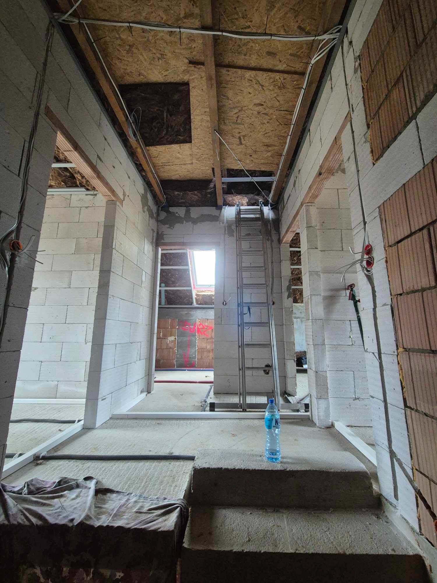 Wnętrze w trakcie budowy: surowe ściany z betonu komórkowego, widoczne przewody elektryczne pod sufitem z płyt OSB, aluminiowa drabina i butelka wody na schodach.