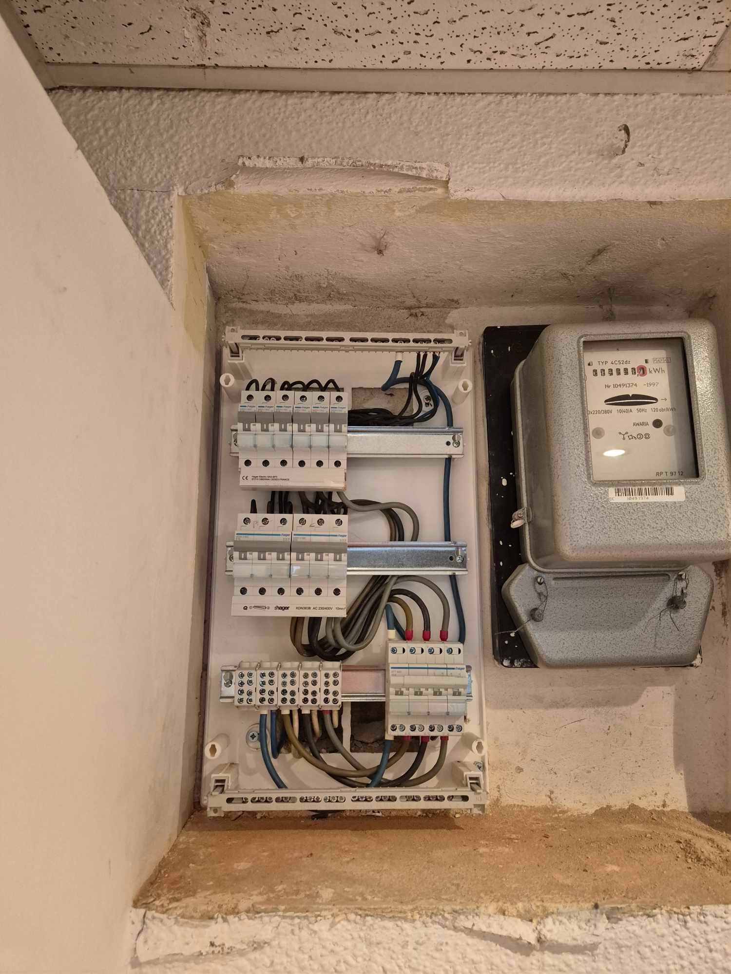 Otwarta skrzynka elektryczna z bezpiecznikami i przewodami, obok stary licznik energii elektrycznej w szarej obudowie, widoczne wnęki w ścianie o nierównej fakturze.