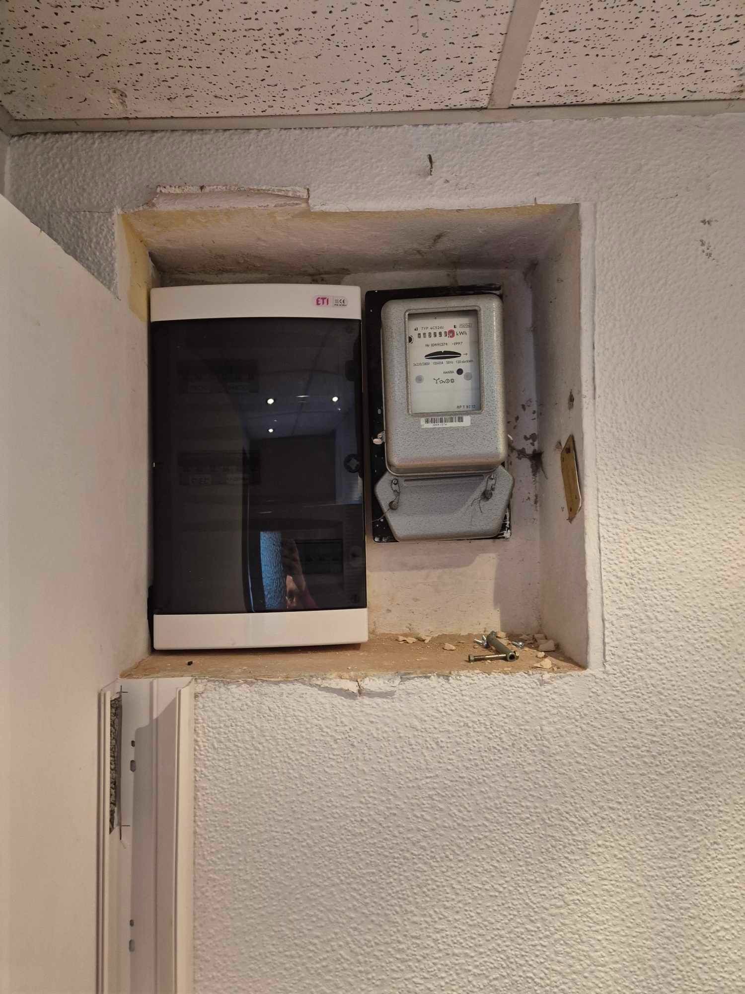 Wnęka w ścianie z zamontowaną skrzynką bezpiecznikową ETI i licznikiem energii elektrycznej, widoczne ślady po wierceniu, surowa ściana z fakturą baranka.