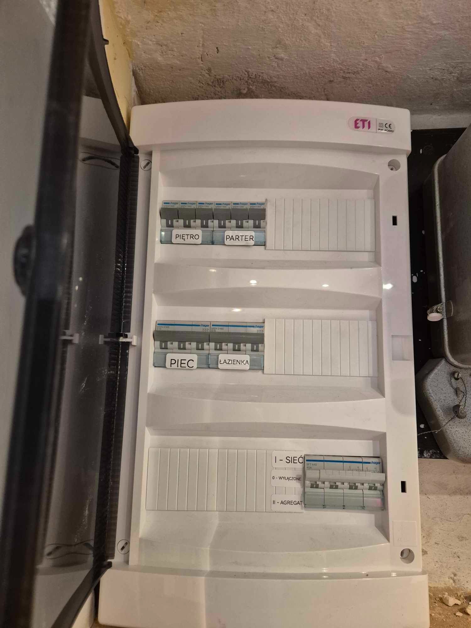 Otwarta, biała skrzynka rozdzielcza z bezpiecznikami, opisane obwody: Piętro, Parter, Piec, Łazienka, Sieć, Agregat, widoczne bezpieczniki marki Hager, zamontowana na ścianie w surowym stanie.