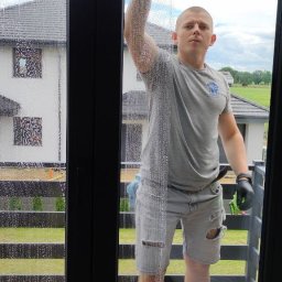 King Cleaning - Mężczyzna w szarych, podartych spodenkach i czarnych rękawicach myje okno za pomocą ściągaczki, widoczny budynek w tle.