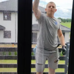 Mężczyzna w szarych, podartych spodenkach i czarnych rękawicach myje okno za pomocą ściągaczki, widoczny budynek w tle.