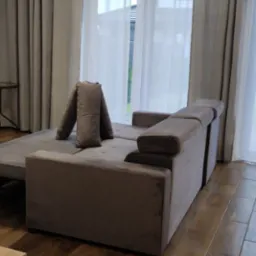 Rozłożona szara kanapa z welurową tapicerką na drewnianej podłodze, poduszki ułożone w nietypowy sposób, w tle jasne zasłony i okno.