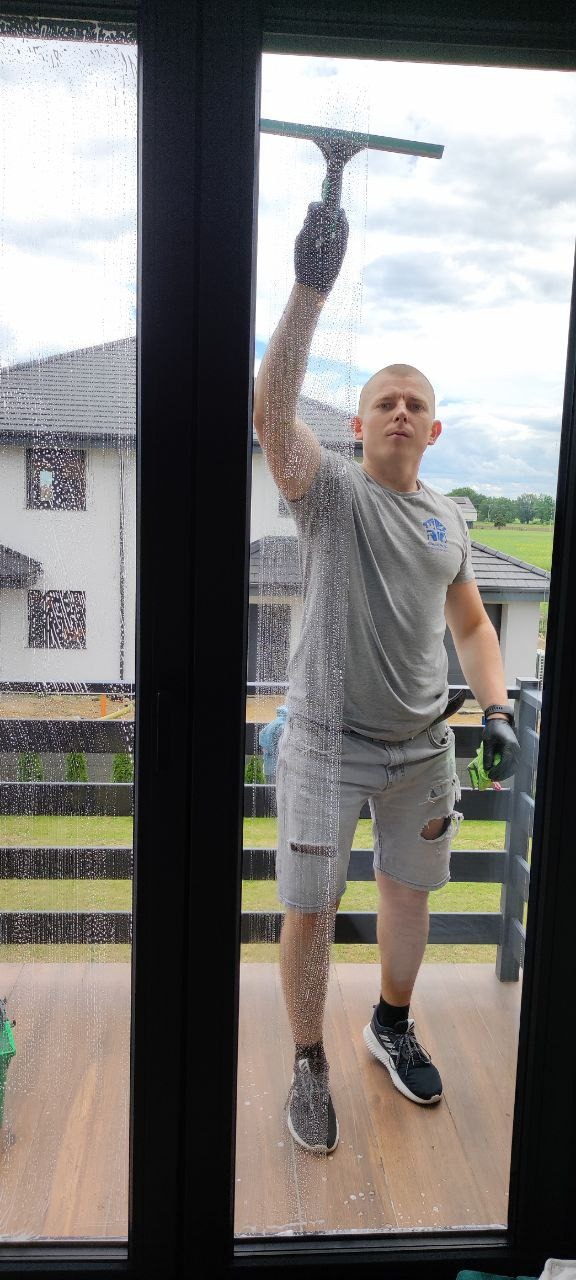 Mężczyzna w szarych, podartych spodenkach i czarnych rękawicach myje okno za pomocą ściągaczki, widoczny budynek w tle.