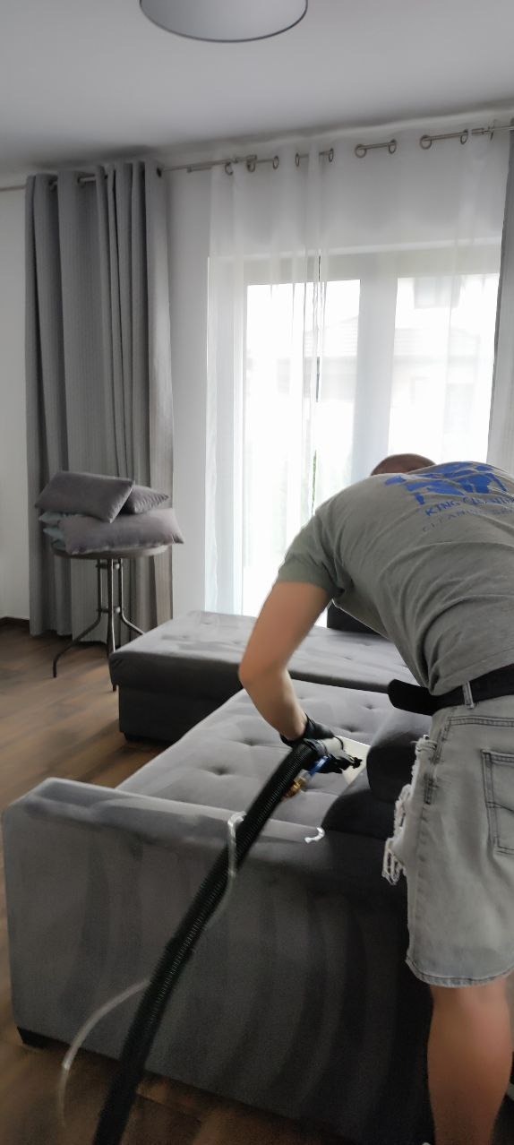 Pracownik w szarej koszulce z logo firmy, w jeansowych spodenkach i czarnych rękawicach czyści szarą, modułową kanapę za pomocą profesjonalnego odkurzacza piorącego z przezroczystą końcówką. W tle...