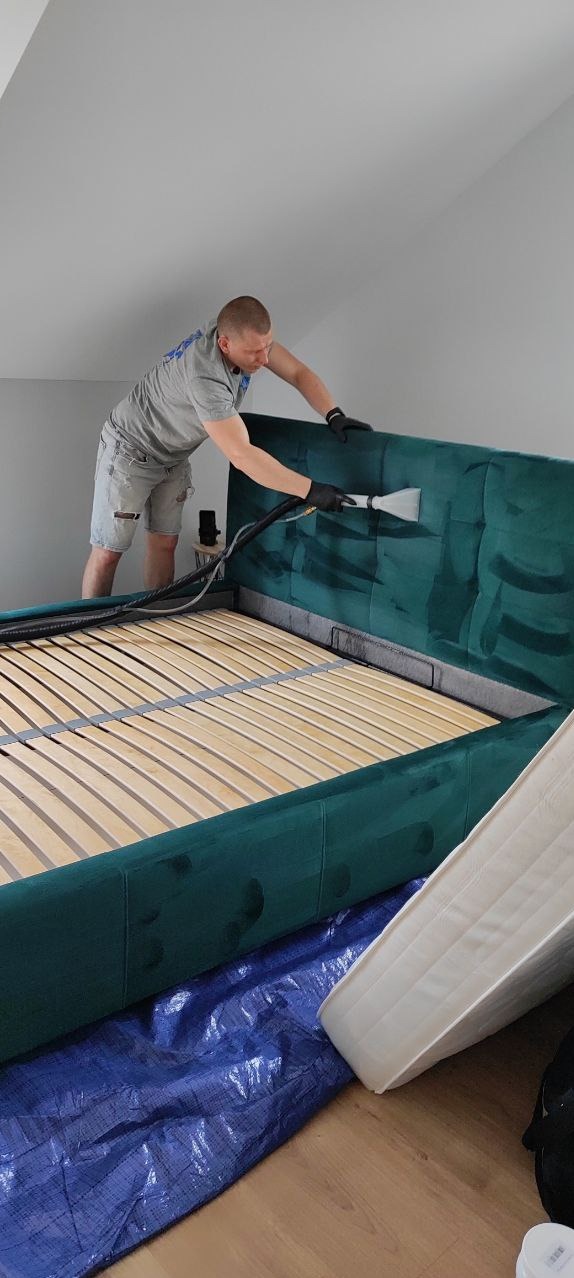 Pracownik w szarym t-shircie i czarnych rękawicach czyści zielony, tapicerowany zagłówek łóżka za pomocą parowego odkurzacza z dyszą. Pod łóżkiem rozłożona niebieska folia ochronna, obok materac.