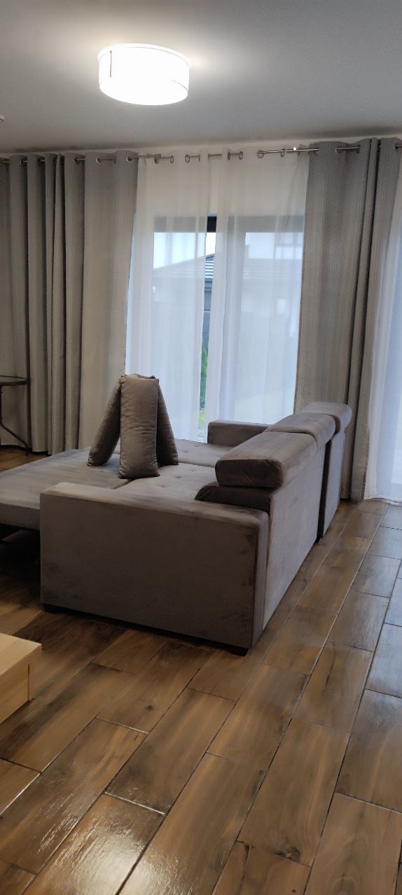 Rozłożona szara kanapa z welurową tapicerką na drewnianej podłodze, poduszki ułożone w nietypowy sposób, w tle jasne zasłony i okno.