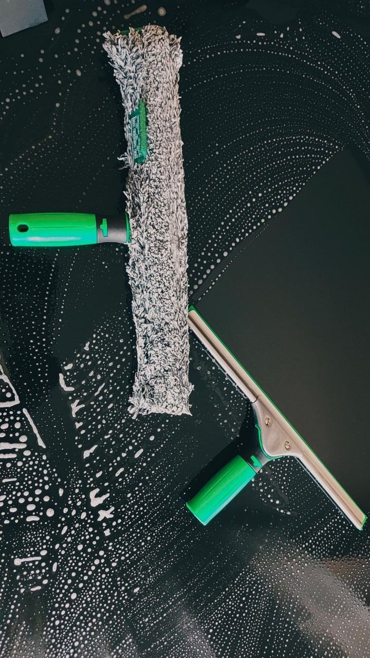 Czyszczona ciemna powierzchnia z widocznymi smugami piany, obok leżą dwa narzędzia do mycia: mop z szarą nakładką i ściągaczka z zielonym uchwytem.