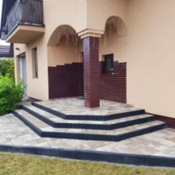 Nowe schody wejściowe do domu, wyłożone beżową kostką brukową z ciemnymi obrzeżami, prowadzące do zadaszonego wejścia z kolumną obłożoną brązowymi płytkami.