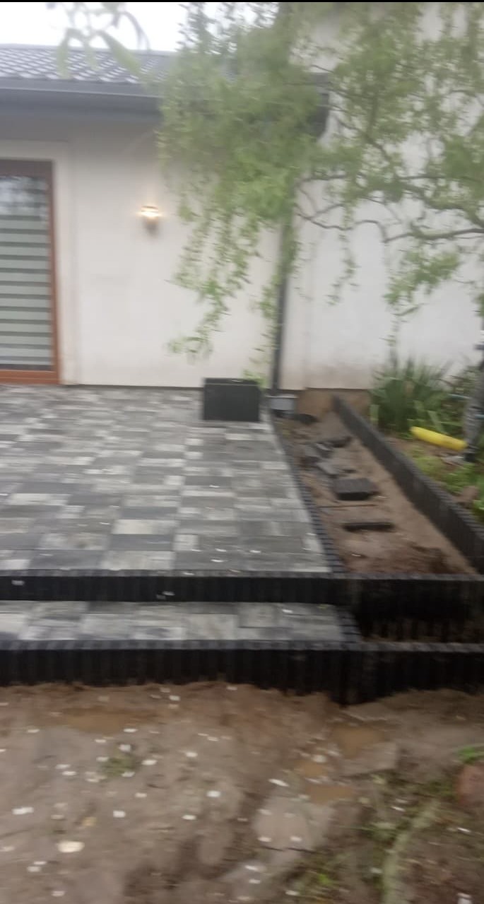 Układanie kostki brukowej w ogrodzie, widoczne fragmenty ułożonej szarej kostki, obrzeża, ziemia i fragment budynku z oknem.