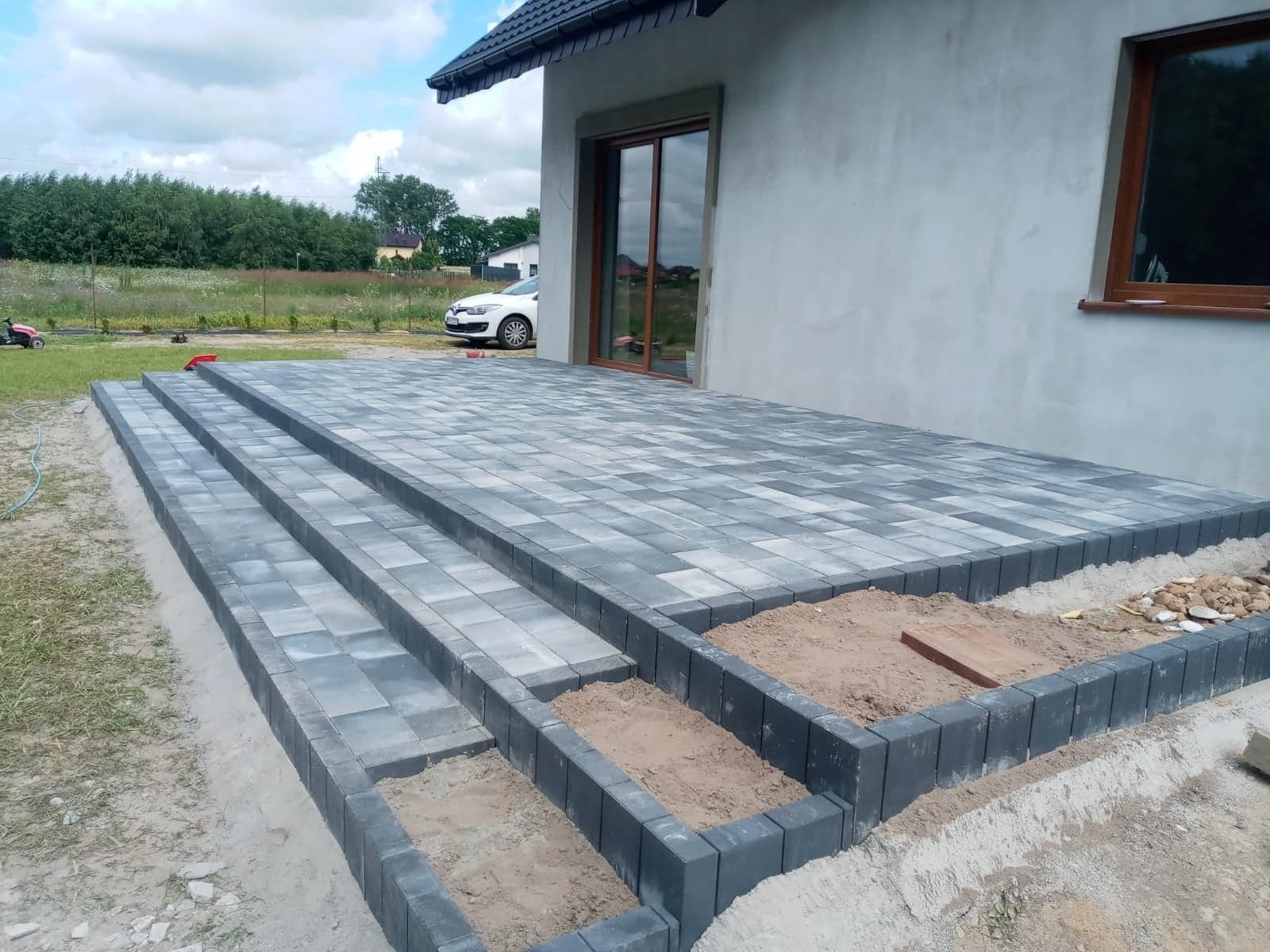 Etap układania szarej kostki brukowej z obrzeżami w Chełmży, widoczne schody i taras przy domu, częściowo zasypane piaskiem.