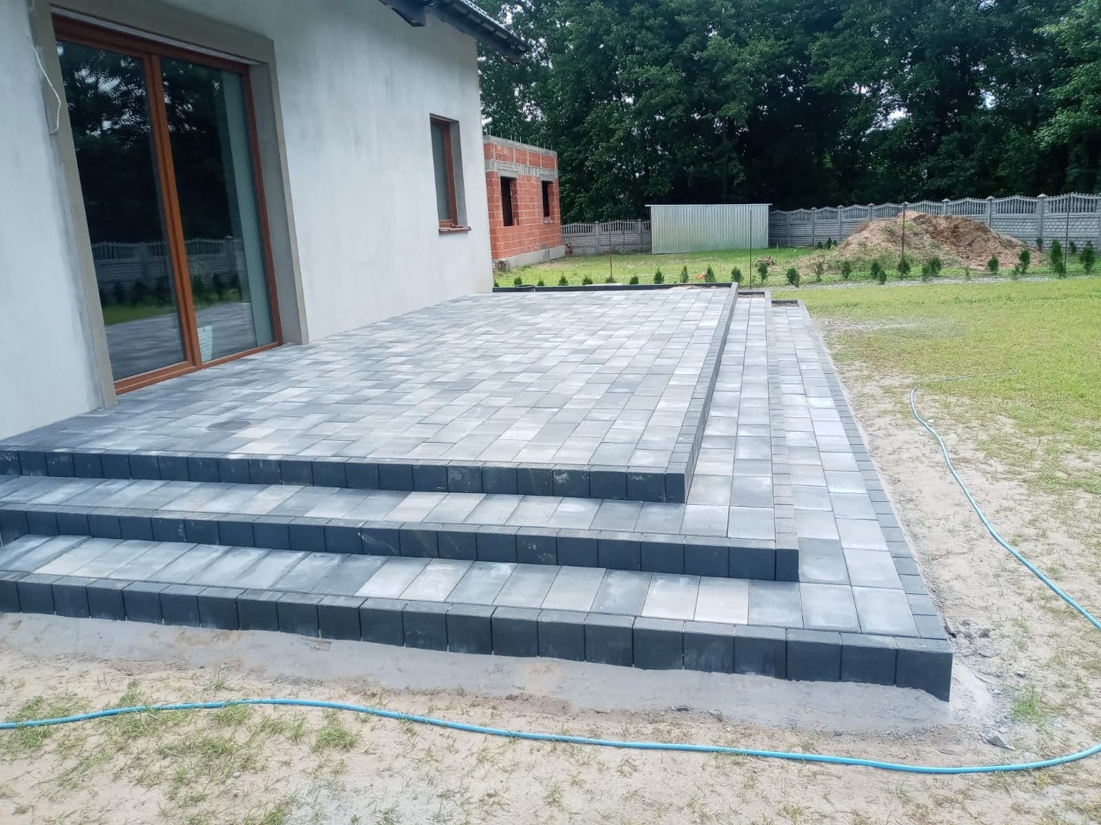 Nowo ułożony taras z kostki brukowej w odcieniach szarości, z trzema stopniami, przylegający do budynku z dużym oknem przesuwnym, widoczne drobne nasadzenia w tle.