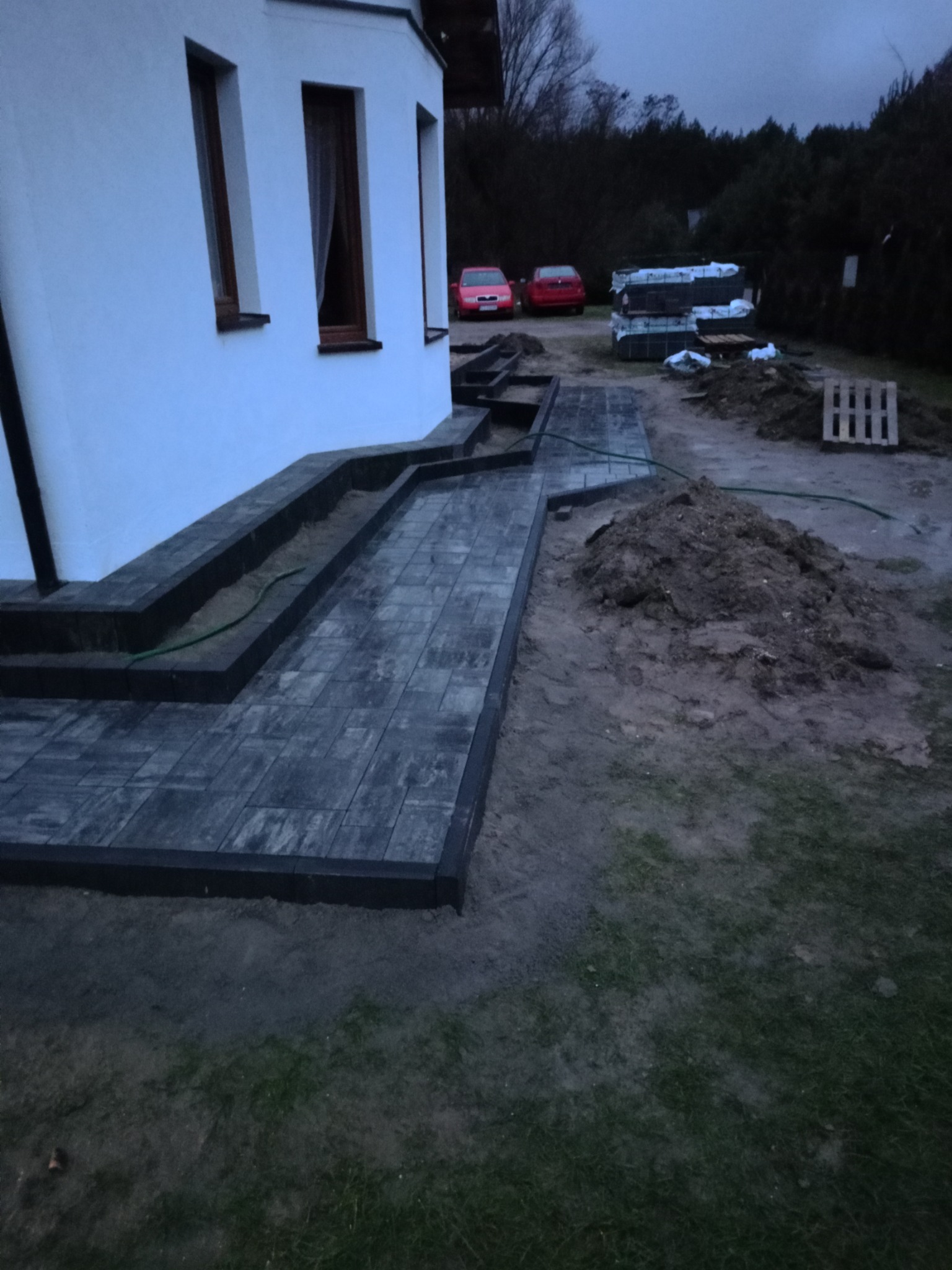 Ułożona ścieżka z ciemnej kostki brukowej prowadząca od schodów domu, widoczny fragment trawnika, hałda ziemi, paleta i zaparkowane dwa czerwone samochody w tle.