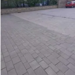 Częściowo wyczyszczona kostka brukowa na podjeździe przed budynkiem z ceglanym murem, widoczna wyraźna różnica w kolorze między umytą a brudną powierzchnią.