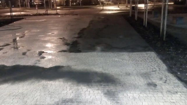 Częściowo umyta kostka brukowa na parkingu w nocy, widoczne mokre i suche obszary, w tle drzewa i oświetlenie uliczne.