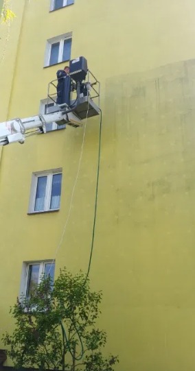 Pracownik na podnośniku koszowym czyści elewację budynku, widoczny fragment umytej ściany, okna i zieleń w dolnej części kadru.