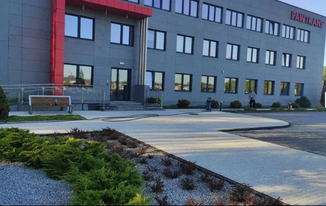 Pracownik w trakcie czyszczenia kostki brukowej przed budynkiem biurowym Pawtrans z użyciem myjki ciśnieniowej. W tle widoczny budynek z szarą elewacją i czerwonymi elementami oraz zadbane rośliny.