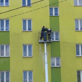 Pracownik na podnośniku koszowym czyści elewację budynku z widocznymi smugami brudu i zaciekami, z użyciem myjki ciśnieniowej.