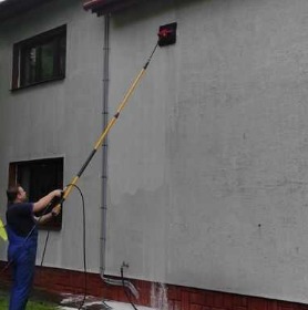 Mężczyzna w niebieskim kombinezonie czyści elewację budynku za pomocą szczotki na długim, teleskopowym kiju. Widać spływającą wodę i pianę.