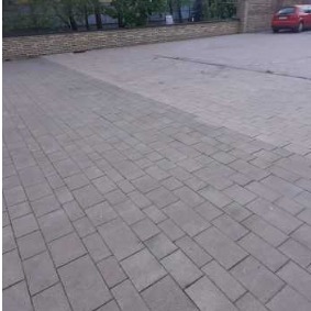 Częściowo wyczyszczona kostka brukowa na podjeździe przed budynkiem z ceglanym murem, widoczna wyraźna różnica w kolorze między umytą a brudną powierzchnią.